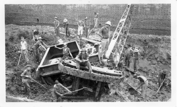 An old-fashioned digging machine with tracks rests on its side in the Fort Pulaski moat. About two dozen men surround and sit on the machine, attempting to dig it out of the mud.